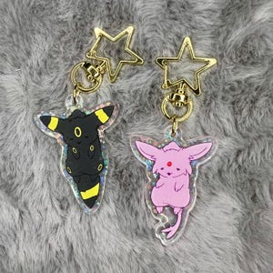 May include: Two keychains featuring Pokemon characters. The keychain on the left features a black and yellow Umbreon with a gold star charm. The keychain on the right features a pink Espeon with a gold star charm.