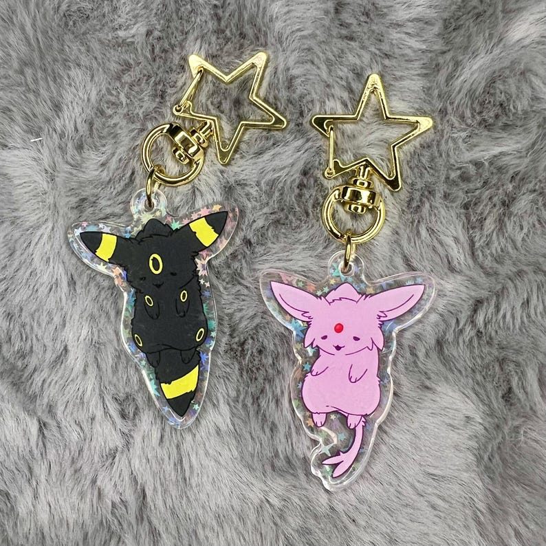 May include: Two keychains featuring Pokemon characters. The keychain on the left features a black and yellow Umbreon with a gold star charm. The keychain on the right features a pink Espeon with a gold star charm.