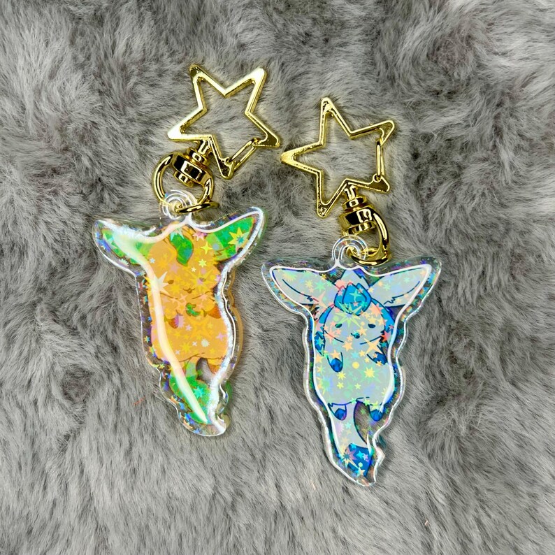 May include: Two iridescent keychains featuring cartoon characters with gold star-shaped charms. The keychain on the left features a yellow character with green accents and the keychain on the right features a blue character with white accents.