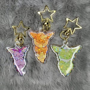 May include: Three keychains featuring different Pokemon characters. Each keychain has a gold star-shaped charm and a clear plastic charm with a glitter background. The Pokemon characters are Eevee, Vaporeon, and Flareon.