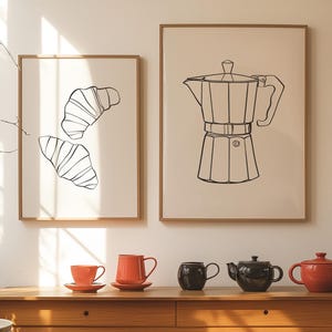 May include: Two framed prints on a white wall, one with a line drawing of croissants, the other with a line drawing of a coffee pot.  A wooden table with four ceramic teapots and mugs in red and black.