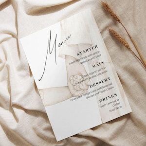 Könnte beinhalten: Eine weiße Menükarte mit dem Wort „Menü“ in eleganter Schrift. Die Speisekarte listet Vorspeisen, Hauptgerichte, Desserts und Getränke auf. Die Speisekarte zeigt eine Aquarellillustration von Händen. Weizenähren sind im Hintergrund.