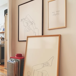 Puede incluir: Tres impresiones artísticas enmarcadas en una pared. Una impresión presenta un dibujo lineal de una persona leyendo un libro, otra dice "one more chapter", y la tercera muestra a una persona leyendo con las piernas levantadas. Un calefactor rojo y plateado está en el suelo.