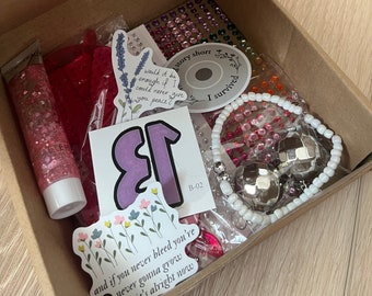 Op de afbeelding: Een kartonnen doos gevuld met verschillende items, waaronder stickers, een roze glitterbuis, een witte kralenarmband met zilveren bedels en een roze en witte sticker met de tekst "and if you never bleed you're never gonna grow and it's alright now".