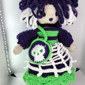 May include: A handmade crochet doll with purple hair and a matching outfit. The doll has a green and purple striped bodice, a white spiderweb skirt, and a small purse with a skull design. The doll's measurements are approximately 8 inches tall.