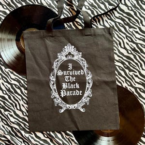 May include: Black tote bag with white text that reads "I Survived The Black Parade" inside a decorative frame. The bag is on a zebra print background.