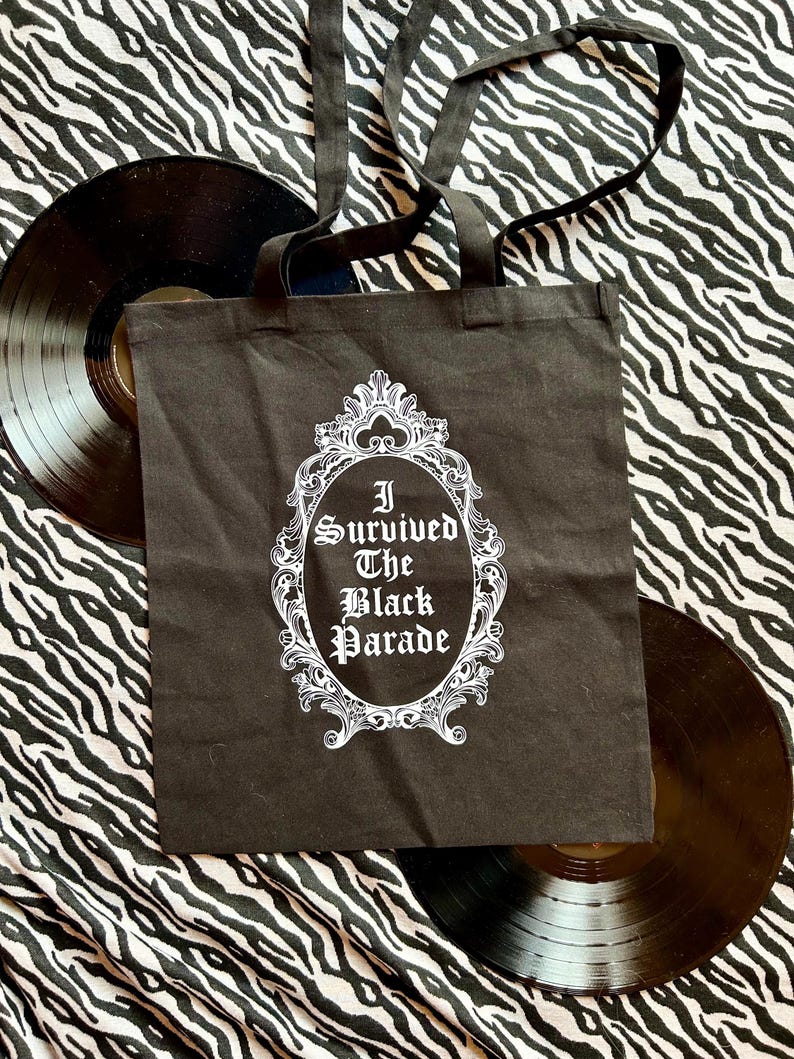 May include: Black tote bag with white text that reads "I Survived The Black Parade" inside a decorative frame. The bag is on a zebra print background.