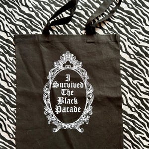 May include: Black canvas tote bag with white text and a decorative frame. The text reads "I Survived The Black Parade".