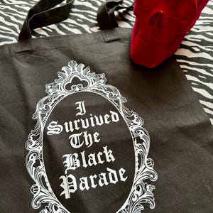 May include: Black canvas tote bag with a white ornate frame and the text "I Survived The Black Parade" inside.