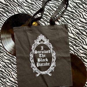 May include: Black tote bag with white text and a decorative frame. The text reads "I Survived The Black Parade".
