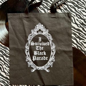May include: Black canvas tote bag with white text and a decorative frame. The text reads "I Survived The Black Parade".