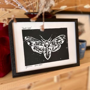 Könnte beinhalten: Ein Schwarzweißdruck eines Totenkopfschwärmers mit einem Schädel auf seinem Thorax. Der Druck ist in Schwarz gerahmt und trägt den Text 'Death moth' und die Unterschrift des Künstlers 'Pixie Iniez' unten rechts.