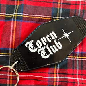 May include: Black coffin-shaped keyring with the words "Coven Club" in white gothic lettering and a white starburst design. The keyring is on a red, black, and yellow tartan background.