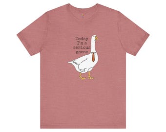 May include: A heathered mauve short-sleeve t-shirt featuring a graphic of a white goose wearing a thin orange necktie. The text "Today I'm a serious goose" is printed in black typewriter-style font next to the goose, which has yellow feet and a yellow beak.