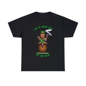 Puede incluir: Camiseta negra con un diseño de duende de dibujos animados. El duende, con pelo rojo y sombrero verde, está sentado en una olla de oro. El texto "I'M A WEE BIT H'IRISH" está impreso en verde encima y debajo de la imagen.