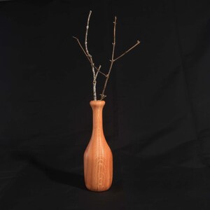May include: A wooden vase in a bottle shape, with a light brown hue and visible wood grain. The vase contains several thin, bare branches. The background is a solid black.