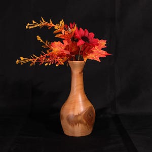 May include: A wooden vase with a light brown finish, holding a bouquet of fall-colored artificial flowers. The arrangement includes red sunflowers, orange maple leaves, and yellow sprigs. The vase has a rounded base and a slender neck.
