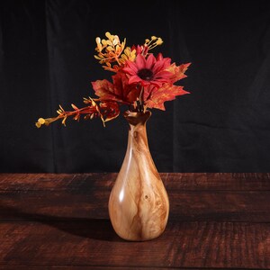May include: A wooden vase with a teardrop shape holds a vibrant arrangement of artificial flowers. The bouquet includes a red sunflower, maple leaves, and yellow sprigs. The vase is made of light brown wood with a natural grain pattern, set against a dark background.