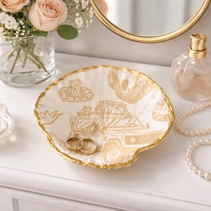 May include: A white, shell-shaped trinket dish with a gold rim and gold illustrations of a vehicle, campfire, and other outdoor elements. Two gold rings rest inside the dish. A perfume bottle and jewelry are in the background.