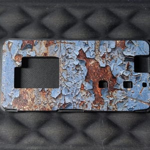 May include: A rectangular phone case featuring a weathered, distressed finish with a pattern of peeling light blue paint revealing a dark, textured rust-colored surface underneath. The case includes precise cutouts for a camera and buttons.