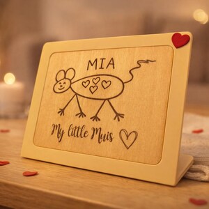 Può includere: Una cornice in legno con un disegno di un topo dei cartoni animati e il nome "MIA". Il testo "My little Mouse" è sotto il disegno. Un piccolo cuore rosso è attaccato all'angolo superiore. La cornice è su una superficie di legno.