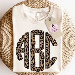 May include: Cream-colored t-shirt with a leopard print monogram of the letters ABC. The letters are outlined in black and filled with a gold glitter effect. A tag with text is attached to the shirt.
