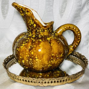 May include: A ceramic pitcher with a rounded, bulbous body and a high-arched spout, featuring a glossy brown glaze with yellow speckled accents. The handle has a beaded texture along its outer edge, and the interior of the pitcher is white.