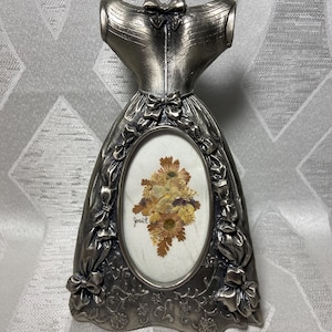 Pode incluir: Uma moldura prateada em forma de vestido com uma abertura oval. A moldura é adornada com laços e detalhes florais. A moldura oval contém flores secas e a assinatura "Joan E".