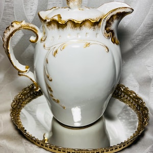Puede incluir: Una jarra de porcelana blanca con tapa, que presenta volutas pintadas en oro y bordes festoneados. La jarra tiene un asa curva y se asienta sobre una bandeja de espejo redonda con borde dorado y un diseño de metal perforado.
