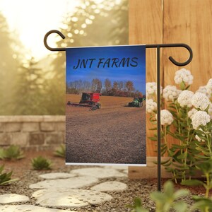 Könnte beinhalten: Eine Gartenflagge mit blauem Himmel als Hintergrund und einem roten und grünen Traktor auf einem Feld. Die Flagge trägt den Schriftzug "JNT FARMS" in schwarzer Schrift.