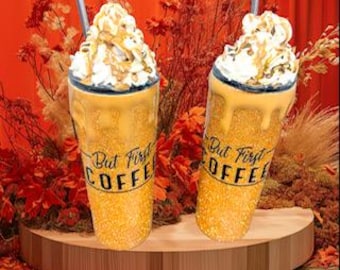 Könnte beinhalten: Zwei goldene Glitzerbecher mit schwarzen Deckeln und schwarzen Strohhalmen. Die Becher sind mit weißer Schlagsahne und einem Karamell-Drizzle verziert und tragen den Schriftzug "But First Coffee" in schwarzer Schrift.