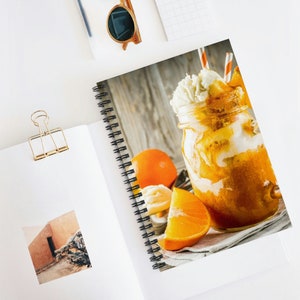 Può includere: Quaderno a spirale con un'immagine vibrante di un dessert all'arancia a strati in un barattolo di vetro, sormontato da panna montata e una cannuccia. Una fetta d'arancia e un'arancia intera sono accanto al barattolo. Il quaderno è aperto su una superficie bianca.