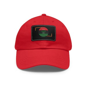 Op de afbeelding: Een rode baseballpet met een gebogen klep. Een zwart rechthoekig embleem op de voorkant toont een cirkelvormig ontwerp met de tekst "1865" en het woord "OFFICIAL". Het ontwerp bevat rode, groene en zwarte kleuren.