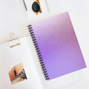 May include: A spiral-bound notebook with a purple glitter gradient cover. The notebook is open, revealing blank white pages. A gold binder clip, sunglasses, and a small notepad are also visible.