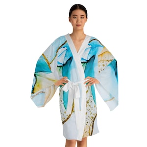 Puede incluir: Bata blanca estilo kimono con un diseño de acuarela en tonos azules, turquesas y dorados. La bata tiene cuello en V y cinturón blanco anudado. Las mangas son anchas y fluidas, y el estilo general es elegante y artístico.