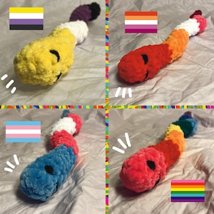May include: Four crocheted worm plushies, each with a different color scheme representing a pride flag. The worms have a smiling face and are lying on a white surface. The flags are: yellow, black, and white; red, orange, and yellow; light blue, white, and pink; and red, orange, yellow, green, blue, and purple.