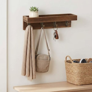 Peut inclure: Une étagère murale en bois brun foncé dotée de quatre crochets en métal. L'étagère supporte une petite plante succulente en pot. Une écharpe en tricot beige, un petit sac bandoulière beige et des clés sur un porte-clés en cuir brun y sont suspendus.