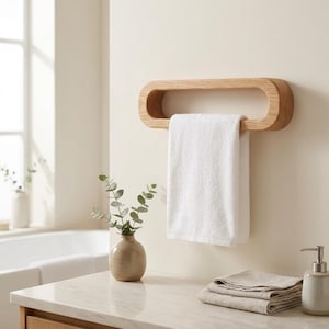 Peut inclure: Un porte-serviettes en bois clair de forme ovale allongée fixé au mur. Une serviette blanche unie est suspendue à travers l'ouverture centrale du cadre en bois. Le grain du bois est visible, révélant une texture naturelle et une teinte brune.