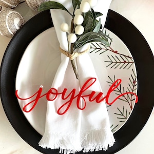 May include: A white napkin with a sprig of white berries and green leaves tied around it. The napkin is placed on a black and white plate with a red wooden sign that says "joyful".