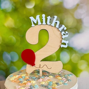 May include: Wooden cake topper with the number 2, the name "Mithran" and a red balloon. The topper is on a white cake with colorful sprinkles.