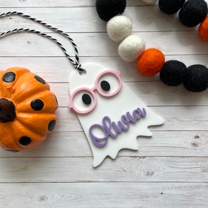 Pu&ograve; includere: Ciondolo bianco a forma di fantasma con occhiali rosa e il nome "Olivia" scritto in viola. Il ciondolo &egrave; appeso a uno spago nero e bianco. Il ciondolo &egrave; su uno sfondo di legno bianco con una piccola zucca arancione con puntini neri.
