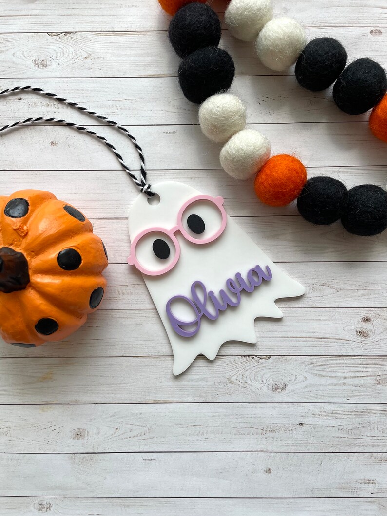 Pu&ograve; includere: Ciondolo bianco a forma di fantasma con occhiali rosa e il nome "Olivia" scritto in viola. Il ciondolo &egrave; appeso a uno spago nero e bianco. Il ciondolo &egrave; su uno sfondo di legno bianco con una piccola zucca arancione con puntini neri.