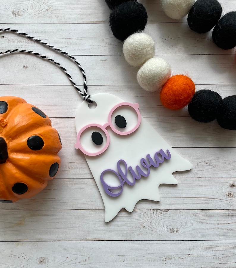 Pu&ograve; includere: Etichetta di Halloween a forma di fantasma bianca con occhiali rosa e il nome Olivia scritto in viola. L'etichetta &egrave; appesa a uno spago bianco e nero.
