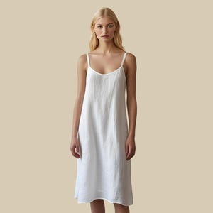 May include: A white, knee-length slip dress with thin straps. The dress has a simple, flowing design and is made of a lightweight fabric. The model is standing in front of a neutral background.