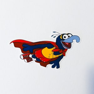 May include: A cartoon image of a blue character with a long, light blue nose, wearing a red cape and red shoes. The character has a yellow vest with gold buttons and a red neckerchief. The character is flying with its arms outstretched.