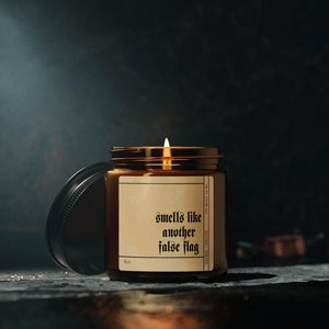 May include: A brown glass candle jar with a black lid. The candle is lit and the label reads "smells like another false flag".