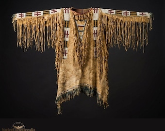 Puede incluir: Una camisa de piel de ante color canela con largos flecos a lo largo de las mangas y el dobladillo. La prenda está decorada con patrones geométricos de cuentas blancas, rojas y negras en hombros, pecho y mangas, con un detalle de cordones.