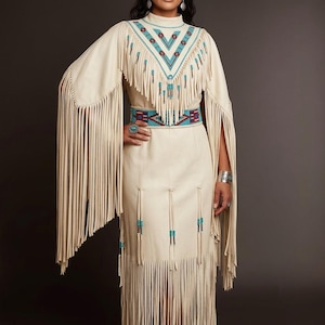 May include: A cream-colored dress featuring long, flowing fringe along the sleeves and hem. The bodice and waist are adorned with intricate geometric beadwork in shades of turquoise, red, and dark blue. The garment has a high neckline.