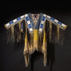 May include: A Native American-style garment with a blue, yellow, and tan color scheme. The top features beaded accents in white and blue, with long fringe detailing in brown and tan. The garment has a V-neck and a rectangular shape.