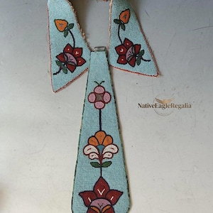 Op de afbeelding: Een lichtblauwe kraag en stropdas set met kralen en bloemmotieven in rood, oranje, roze en groen. Het ontwerp bestaat uit een puntige kraag en een lange stropdas, beide versierd met ingewikkeld kralenwerk. De tekst NativeEagleRegalia is zichtbaar.